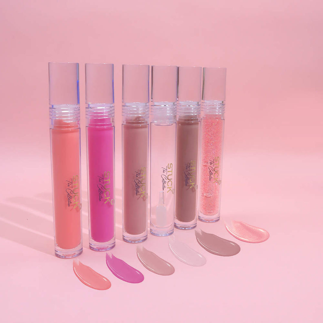 Five tubes of lip gloss from Jazz Beauty Supply in Detroit stand upright on a pink surface, each in pink, purple, nude, brown, and glitter shades with matching swatches displayed against a vibrant pink background.