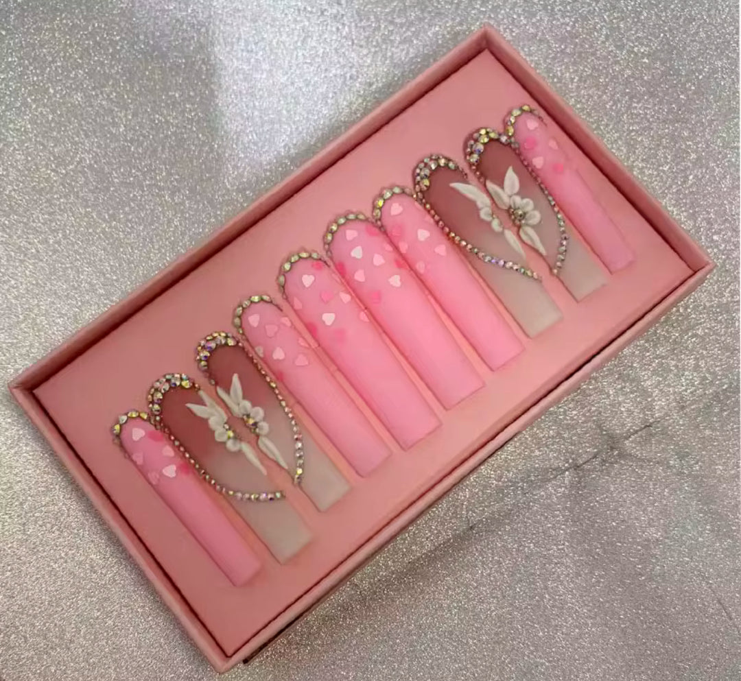 A box of long, pink press-on nails with heart and butterfly designs, adorned with small rhinestones and gold accents, displayed on a sparkly surface—available at Jazz Beauty Supply in Detroit.