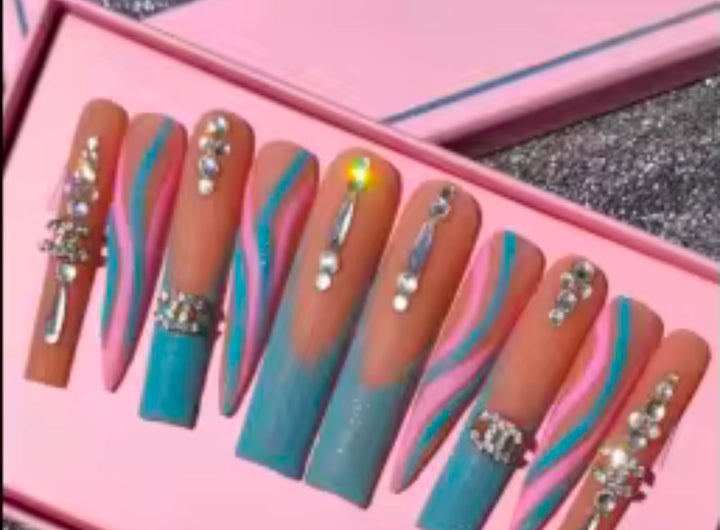 A set of long false nails with a pink and blue wavy design, decorated with rhinestones and metallic charms, displayed in a pink box—available at Jazz Beauty Supply in Detroit.
